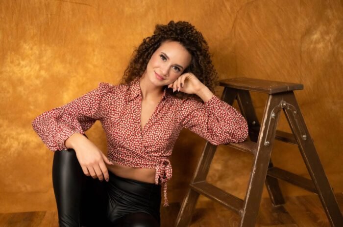 Portré fotózás Debrecenben - A képen egy nő látható, aki egy létrára támaszkodva mosolyog. Pirossal és fehérrel mintás blúzt és fekete bőrnadrágot visel. A háttér narancssárga színű, amely kontrasztot ad a női karakter éles vonásainak és kedves mosolygásának. A fotó barátságos, természetes hangulatú.