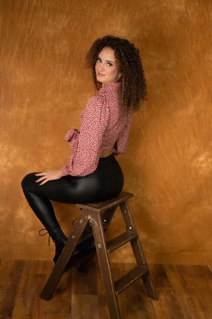 A képen egy nő ül egy létrán, miközben hátrafordul és mosolyog a kamerába. Fekete bőrnadrágot és piros-fehér mintás, laza blúzt visel. A háttér narancssárga árnyalatú, ami meleg hangulatot ad a fényképnek. A színek és a lágy fények kiemelik a nő természetes szépségét.