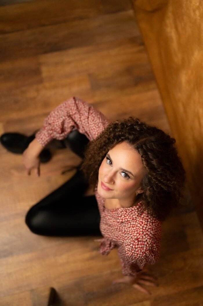 Portré fotózás Debrecen - A képen egy nő ül, fekete bőrnadrágban és egy virágos felsőben, miközben a kamerába néz és kedvesen mosolyog. Az enyhén felülről készült felvételen a haját lágy göndör loknik keretezik, ami a fényekkel és a háttérben lévő meleg színekkel egyaránt harmonizál.