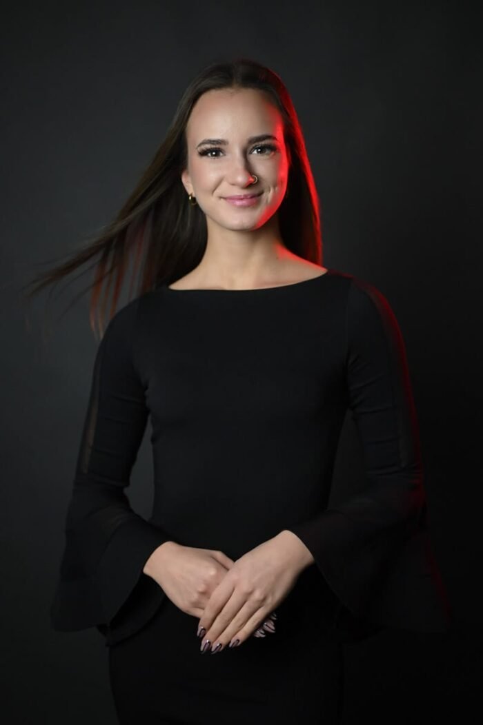 Fiatal nő elegáns fekete ruhában áll, kezét finoman összekulcsolva, piros fényhatással kiemelve, sötét stúdió háttér előtt Debrecenben.