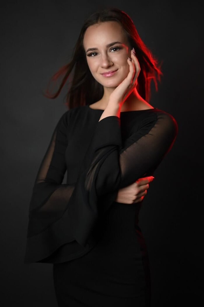 Fiatal nő fekete ruhában pózol, kezét az arcához emelve, piros fényhatással kiemelve, sötét stúdió háttér előtt Debrecenben, finom mosollyal.