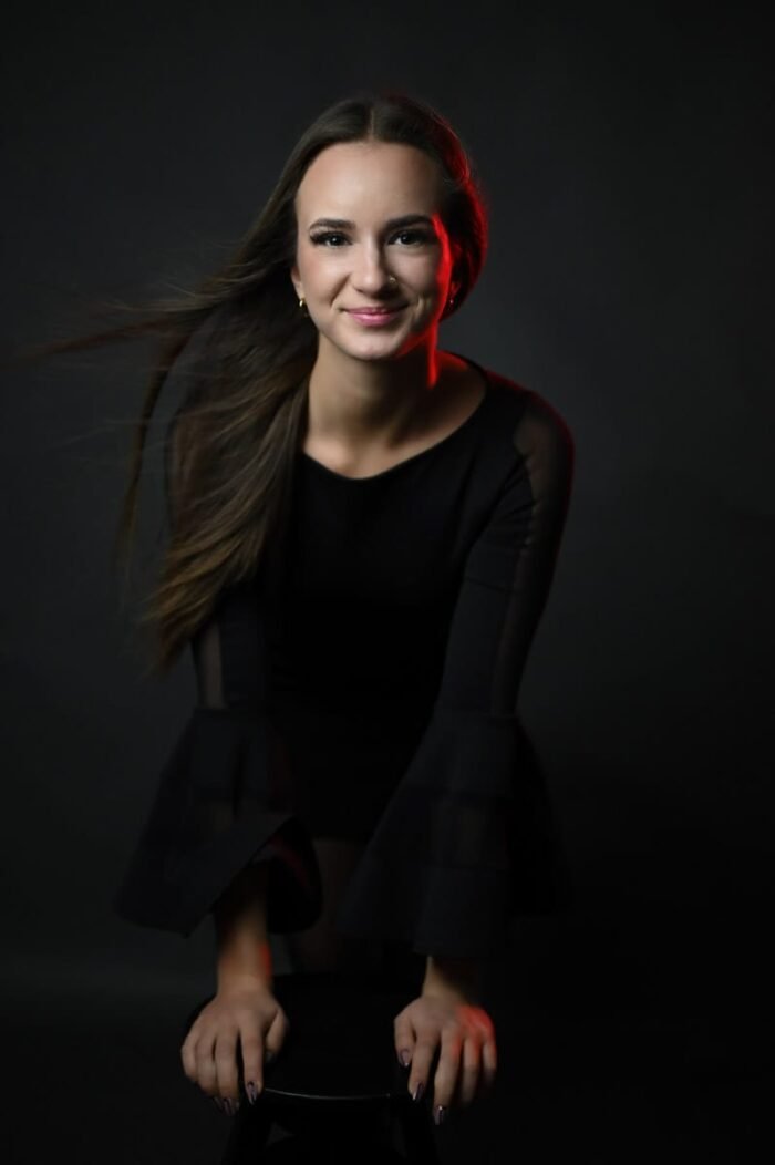 Debreceni fotós, Fiatal nő fekete ruhában pózol egy széken előrehajolva, piros fényhatással és finoman mozgó hajjal, drámai megvilágításban, sötét háttér előtt.