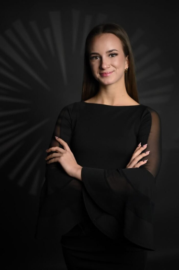 Portré fotó Debrecen. Fiatal nő fekete ruhában áll karba tett kézzel, sugárzó mintázatú kreatív háttér előtt, finom mosollyal, elegáns stílusban.