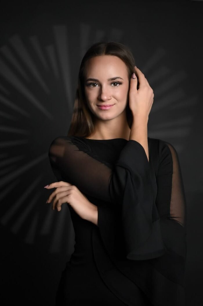 Fiatal nő fekete ruhában áll elegáns pózban, kezét arcához emelve, kreatív sugárzó mintázatú háttér előtt, finom mosollyal.