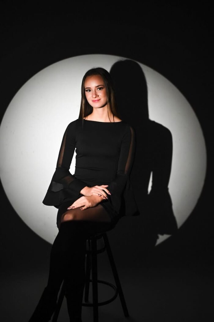 Fiatal nő fekete ruhában ül egy széken reflektorfényben, sötét stúdióban Debrecenben, miközben finom mosollyal néz a kamerába.