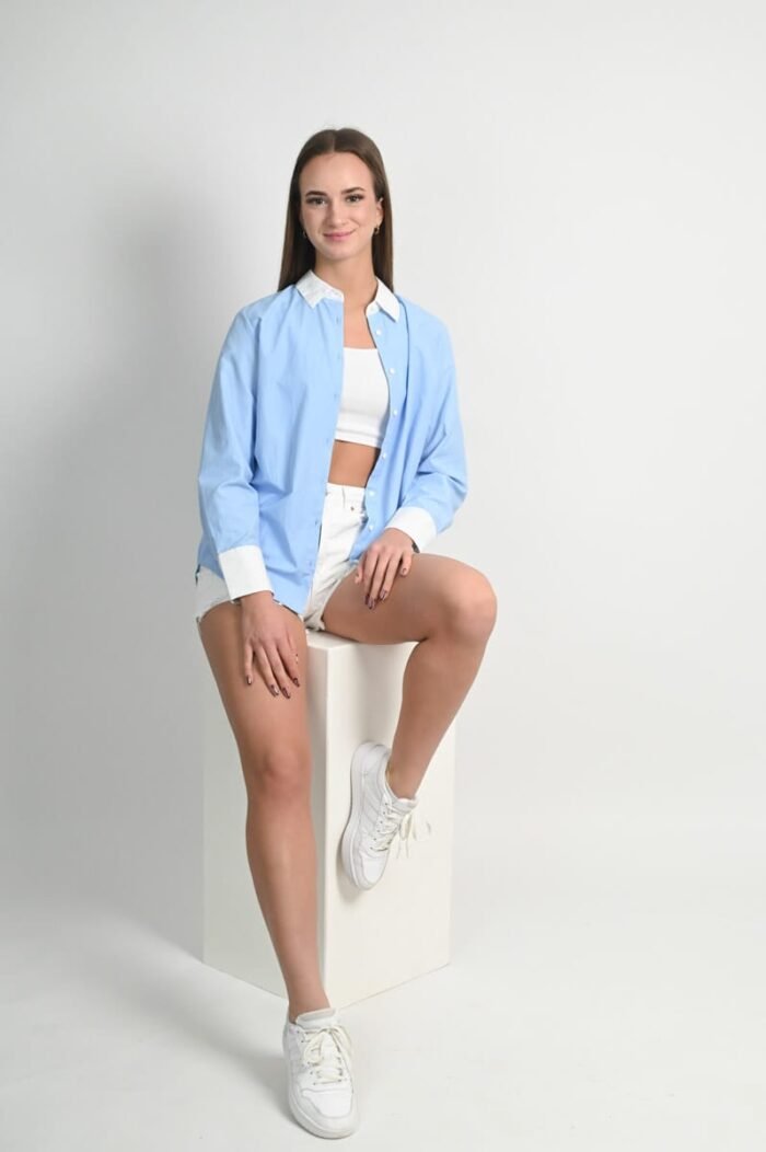 Portré fotó - Debrecen. Fiatal nő kék inget, fehér crop topot és rövidnadrágot viselve ül egy fehér dobozon világos stúdióban, sportcipőben, természetes mosollyal néz a kamerába.