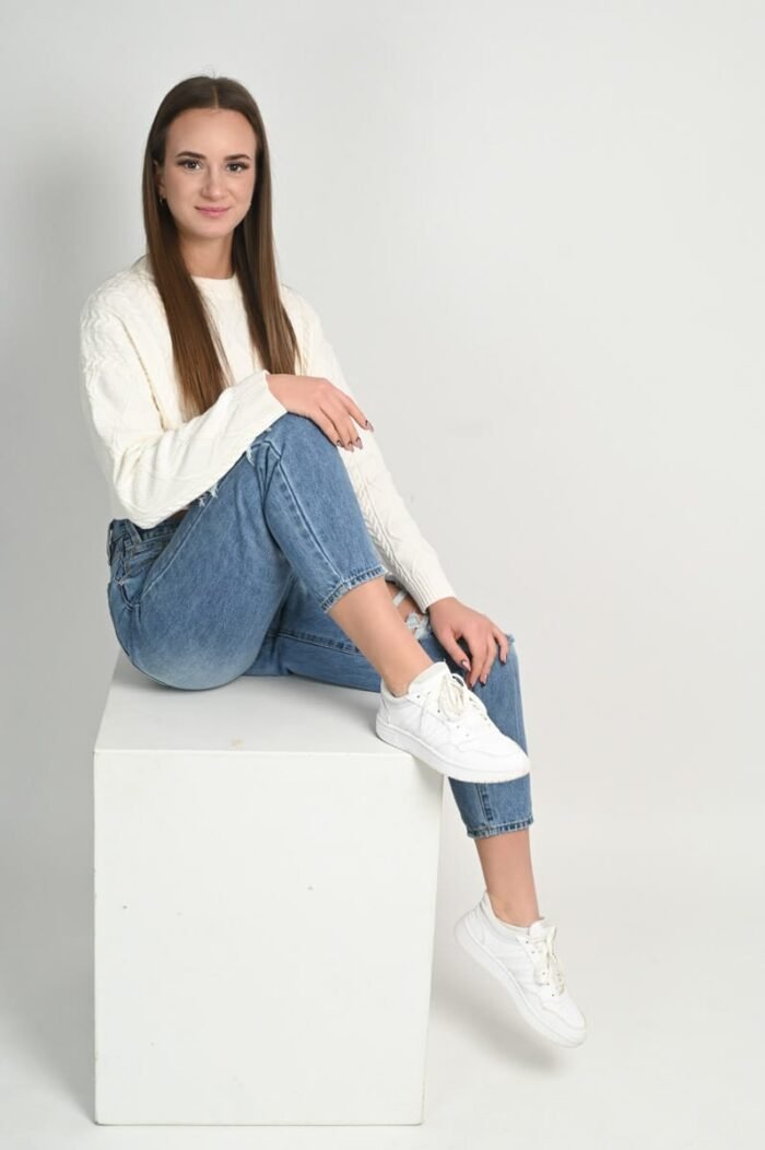 Debreceni fotós. Fiatal nő fehér pulóverben, koptatott farmerben és sportcipőben ül egy fehér dobozon világos stúdióban, miközben természetes mosollyal néz a kamerába.
