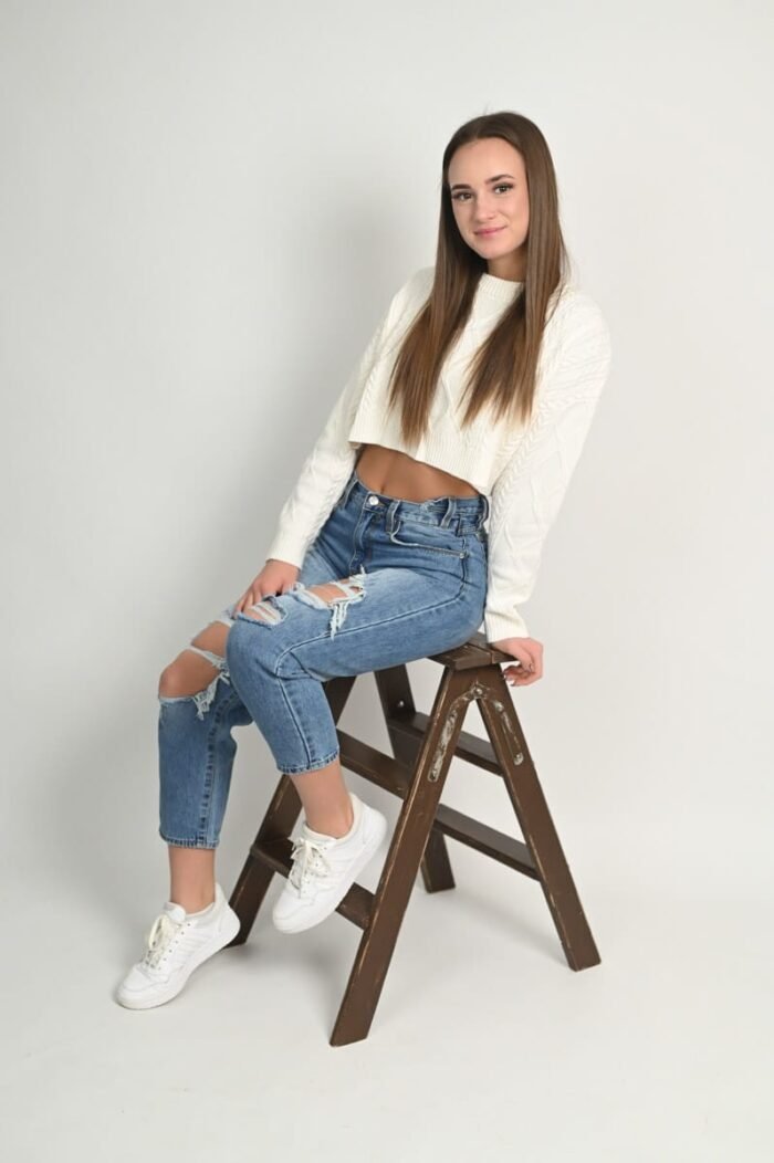 Laza portré fotózás Debrecenben - Fiatal nő fehér pulóverben, koptatott farmerben és sportcipőben ül egy barna fa létrán világos stúdióban, mosolyogva a kamerába.