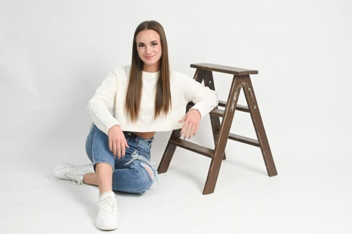Stúdió fotózás Debrecenben.Fiatal nő fehér pulóverben és koptatott farmerben ül a földön világos stúdióban, mellette egy barna fa létra, miközben mosolyogva pózol a kamerának.