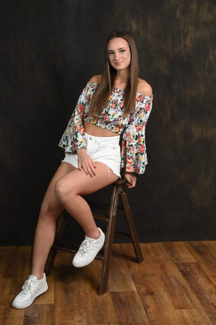 Fiatal nő virágmintás felsőben, fehér rövidnadrágban és sportcipőben ül egy barna széken, természetesen mosolyogva pózol sötét háttér előtt, fa padlón, Debrecen stúdiójában.