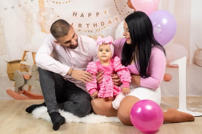 Egy boldog család közösen ünnepli a kislány első születésnapját. Az anya és az apa mosolyogva néznek a kislányukra, miközben a kicsi, rózsaszín ruhában és fejpántban boldogan ül. A háttérben a "Happy Birthday" felirat, lufik és egy nagy "1" szám emeli a szülinapi hangulatot