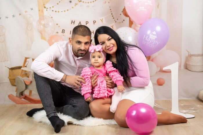 Egy boldog család három tagja közösen ünnepli a kislány első születésnapját. Az anya, az apa és a baba mind rózsaszínben öltöztek, miközben a kislány a szülei között mosolyog. A háttérben színes lufik, egy "Happy Birthday" felirat és a nagy "1" szám látható, melyek tovább emelik a születésnapi hangulatot.