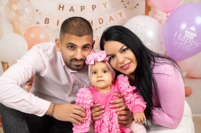 Egy boldog család három tagja közösen örül a kisbaba első születésnapján. Az anya, az apa és a baba mosolygósan ölelkeznek, miközben a kislány rózsaszín ruhájában és hajpántjában figyel a kamerába. A háttérben színes lufik és "Happy Birthday" feliratú dekorációk láthatók, amik tovább fokozzák az ünnepi hangulatot.