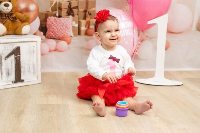 1 éves kislány szülinapi fotózás. Vidám kisbaba piros szoknyában és 1-es számú díszítéssel, rózsaszín lufikkal és ajándékdobozokkal körülvéve, születésnapi fotózás során.