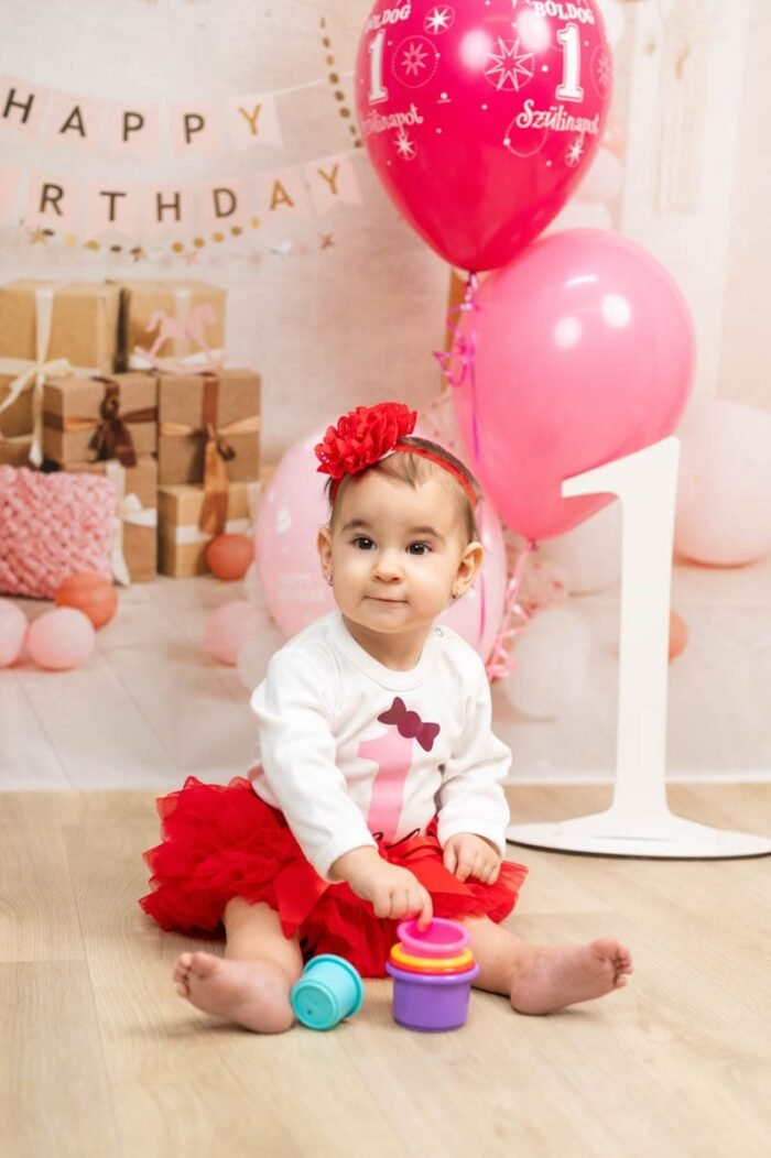 Kisgyermek piros szoknyában, rózsaszín és piros lufikkal, ajándékdobozokkal és 1-es számú díszítéssel ünnepi háttérben, játékokkal a kezében.