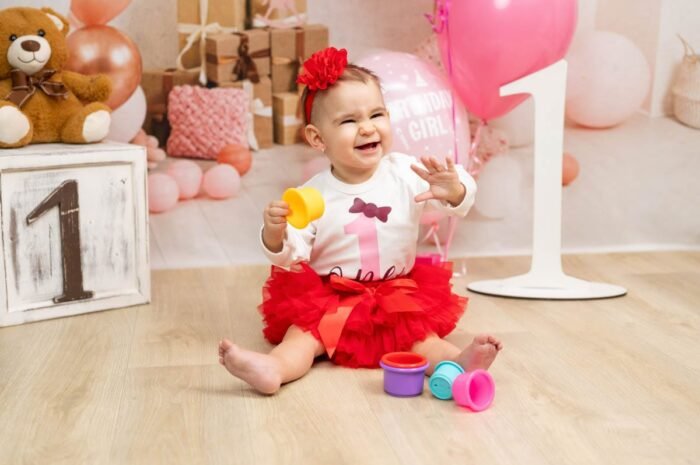 Szülinapi fotózás Debrecenben - Nevető kisbaba piros szoknyában születésnapi dekorációk között, játékokkal a kezében, háttérben lufikkal, plüssmackóval és ajándékdobozokkal.