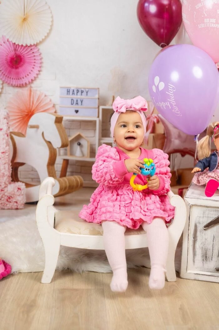 A kis hercegnő boldogan játszik egy színes játékkal, miközben a háttérben színes lufik és dekorációk, köztük a "Boldog Születésnapot" felirat is megjelenik. A kislány rózsaszín ruhában és hajpántban ül egy fehér kis kanapén, miközben mosolygósan néz a kamerába. A baba mellett egy másik plüss játék és díszítés is látható, miközben a fotózáson a születésnap hangulata teljesedik ki.