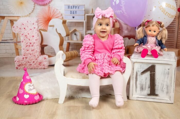 Első szülinapi fotózás - Egy kislány, rózsaszín ruhában és hajpánttal, boldogan ül a fehér kis kanapén a 1. születésnapi fotózáson. A háttérben színes lufik, virágok és egy babát ábrázoló díszítés látható. A kislány mellett egy plüss játék és egy születésnapi sapka is megjelenik, míg a fényképen megjelenik a "Happy Day" és a "Boldog Születésnapot" feliratú dekoráció.