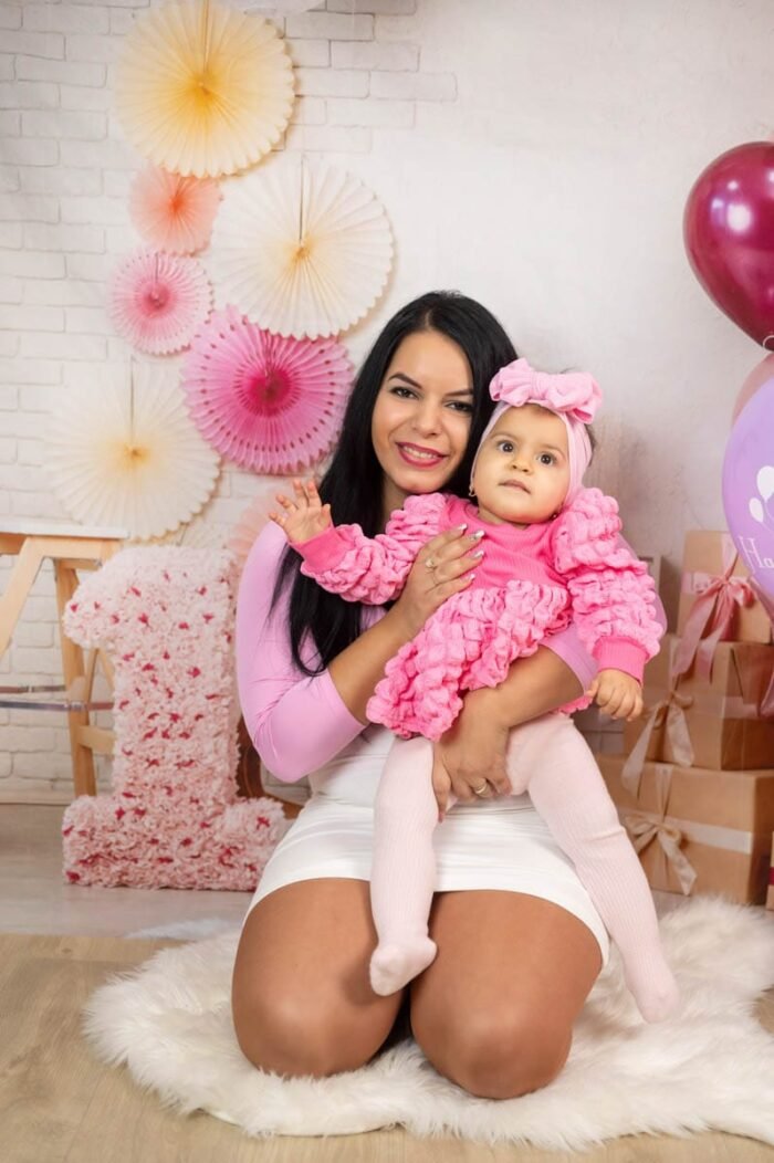 Egy anyuka és kislánya a kislány 1. születésnapján, rózsaszín ruhában és hajpántban. Az anyuka boldogan öleli a kislányát, miközben a háttérben születésnapi dekorációk, ajándékok és lufik láthatók. A kislány boldogan mosolyog, miközben szülei szeretetteljes pillanatait élvezi.