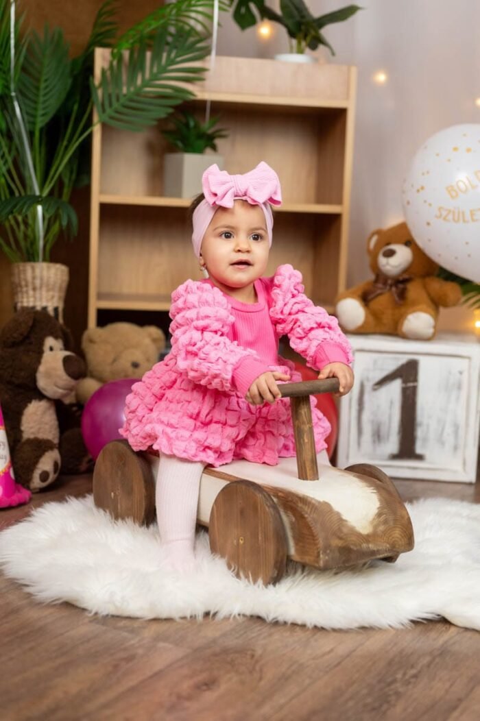 Egy kislány az 1. születésnapján rózsaszín ruhában és hajpántban, miközben egy fa játékautót vezet. A háttérben születésnapi dekorációk, lufik, egy "1"-es szám és egy plüss mackó látható. A kislány boldogan mosolyog, miközben a születésnapi pillanatokat élvezi.