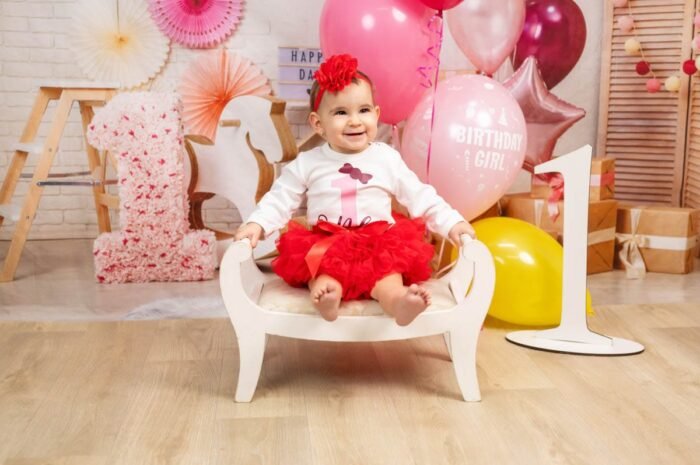 Kisgyermek piros szoknyában, mosolyogva ül egy fehér széken, körülötte születésnapi dekorációk, lufik és egy virágos 1-es díszítés.
