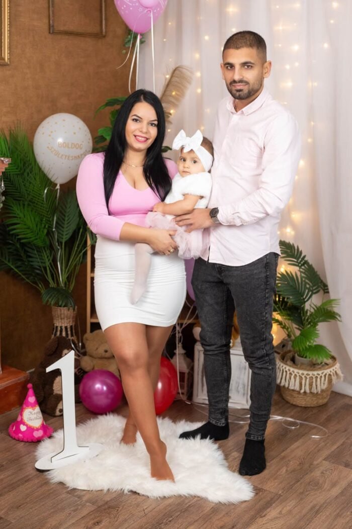 Egy család tagjai egy születésnapi fotózáson, az édesanya, édesapa és a kislány együtt láthatóak. Az anya rózsaszín ruhát visel, míg az apa világos rózsaszín inget. A kislány kis rózsaszín szoknyát és fehér köpenyt visel, fején fehér masnival. A háttérben születésnapi dekorációk és egy nagy "1"-es szám látható.