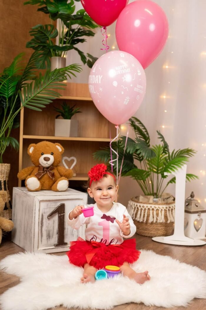 Mosolygós kisbaba születésnapi dekorációval, rózsaszín és piros lufikkal, plüssmackóval, valamint 1-es számú díszítéssel.