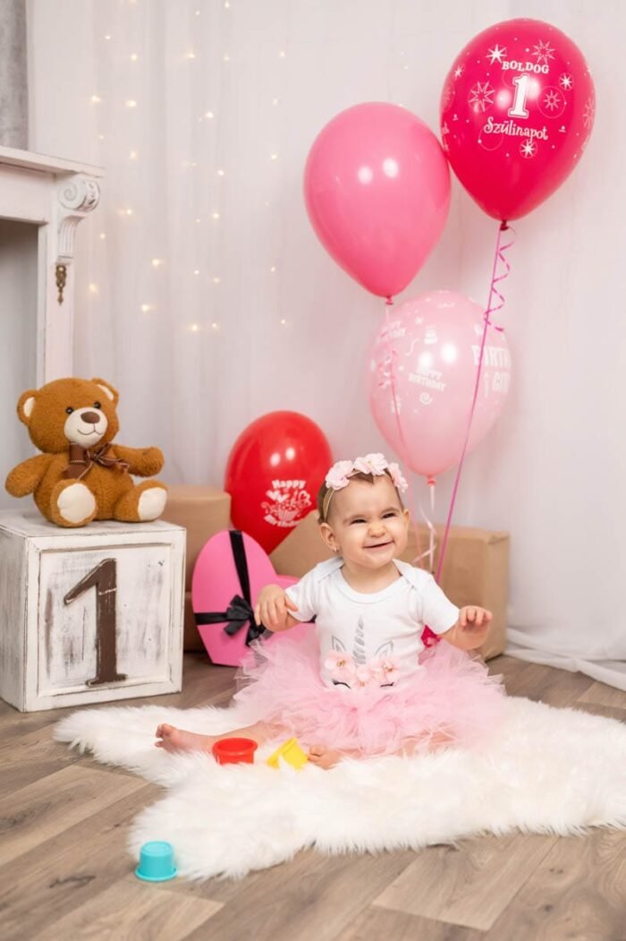 A kis baba boldogan játszik a szülinapi dekorációk között. A háttérben színes ballonok, ajándékok és egy plüssmaci látható. A baba rózsaszín tüll szoknyában és virágos hajpántban ül, miközben színes kúpokat tart a kezében, és boldogan mosolyog. A díszített "1"-es szám és a "Boldog Szülinapot" feliratú ballonok kiegészítik az ünnepi hangulatot.