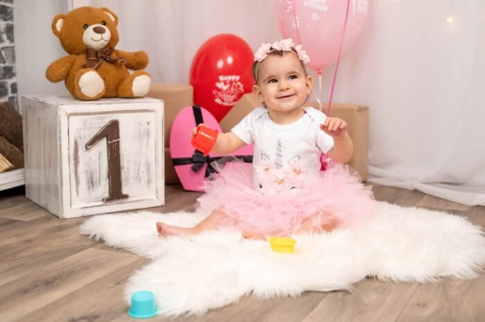 A kis baba boldogan játszik, miközben piros és sárga játékokat tart a kezében. A háttérben színes ballonok és ajándékok láthatók, a kis hercegnő virágos hajpánttal és rózsaszín szoknyával ünnepli első születésnapját. A „1”-es számot ábrázoló dísz is látható a képen.