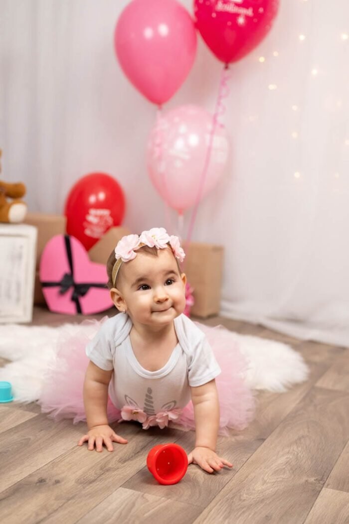 A kis baba boldogan mászik a szőnyegen, miközben egy piros játékkal játszik. A háttérben rózsaszín ballonok és ajándékok láthatóak, a kis hercegnő virágos hajpánttal és rózsaszín szoknyával ünnepli első születésnapját.