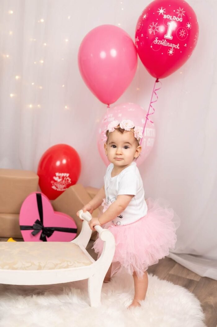 A boldog kis hercegnő egy fehér kis kanapénál támaszkodik, miközben két szép rózsaszín ballon mellett áll. A háttérben ajándékok és díszek láthatók, a kis baba pedig szülinapi hangulatban mosolyog, és a szülei szórakoztató ünnepi pillanatait örökíti meg a fényképezőgép.