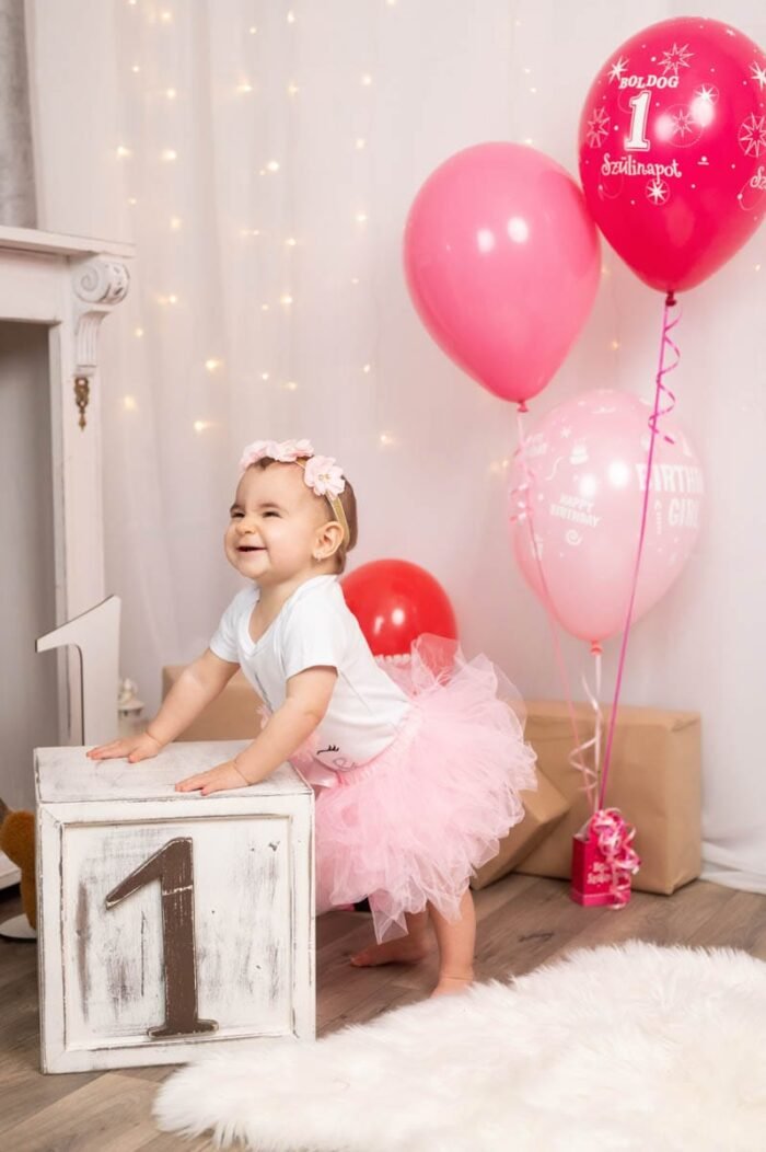Első szülinapi fotózás Debrecenben. A kis hercegnő mosolyogva épp a "1" számú díszhez támaszkodik, miközben ünnepi lufik és ajándékok veszik körül. A háttérben fények és a boldog születésnap hangulatát erősítő díszek láthatók, ahogyan a kislány boldogan játszik a szülinapi pillanatban.
