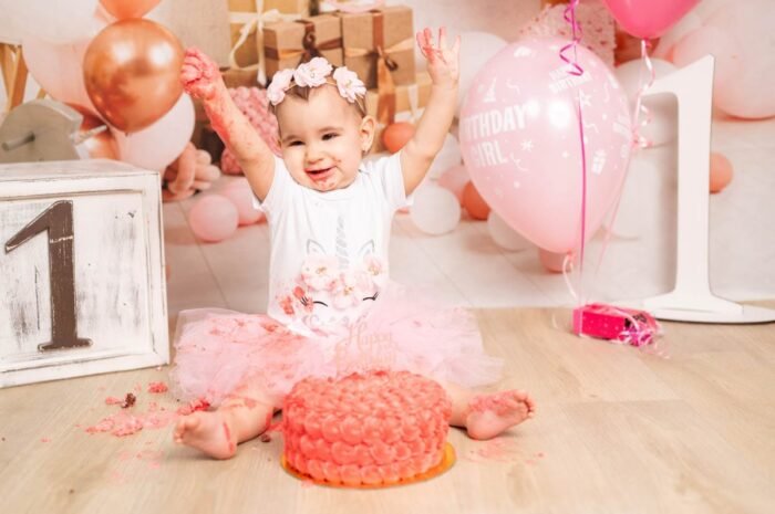 A kislány boldogan ünnepli első szülinapját, miközben teljesen belemerül a torta felfedezésébe. A rózsaszín torta morzsái és krémje az egész területet elfedik, miközben ő boldogan mosolyog. A háttérben lufik és ajándékok adnak ünnepi hangulatot, és a "1"-es számú dísz kiemeli az első szülinapot.