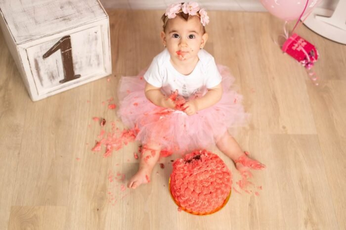 A kislány boldogan felfedezi a születésnapi tortáját, miközben a piros torta morzsái és krémje elborítják a szoknyáját és kezét. A "1"-es számú dísz és a lufik hozzátesznek az ünnepi hangulathoz. Az édes kis virágos fejpánt és a rózsaszín tütü ruha kiemelik a pillanat boldogságát.