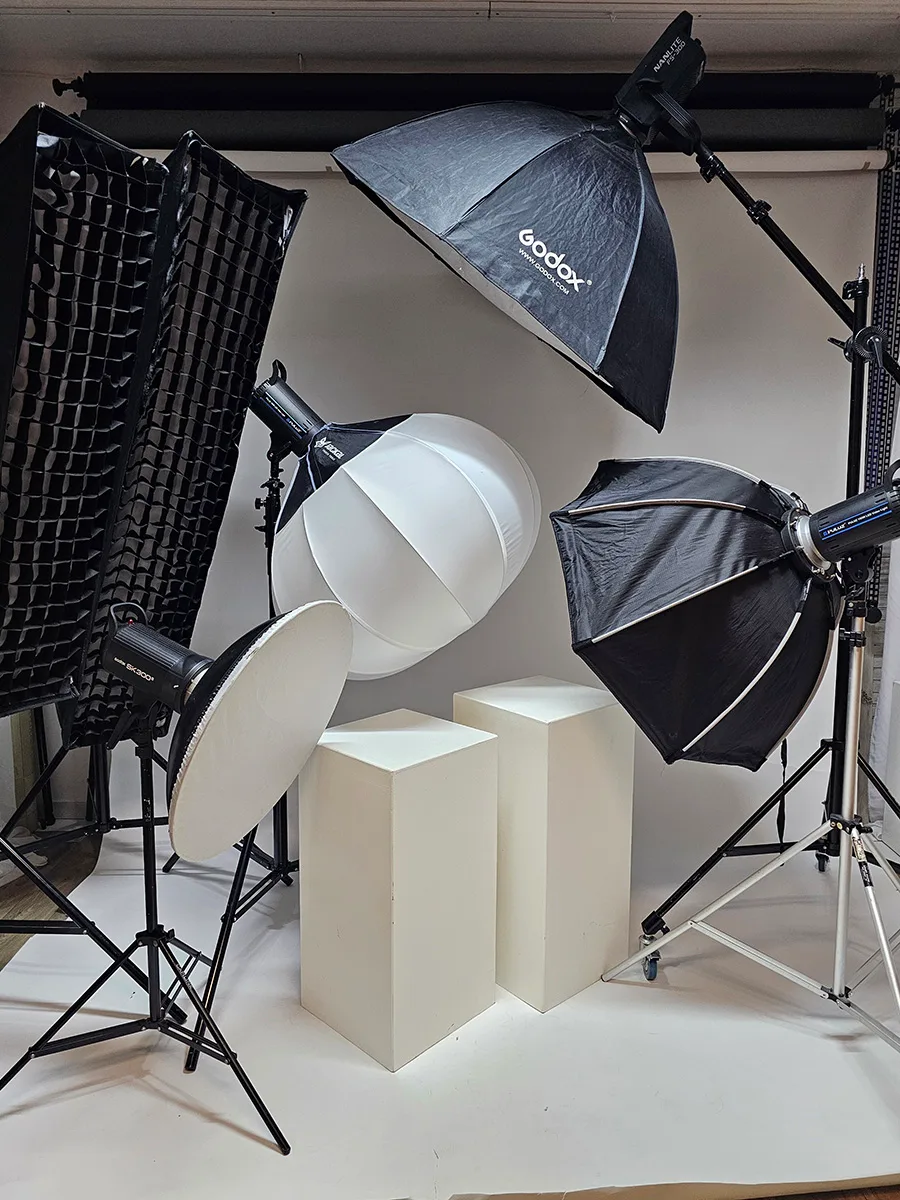 A fotóstúdió bérléséhez készült képen jól láthatóak a professzionális világítóeszközök, mint a Godox vakuk, softboxok, fehér reflexor és különböző lámpák, amelyek lehetővé teszik a precíz fénybeállítást. A stúdióban találhatóak a fehér fotózási bázisok is, amelyek segítenek az egyedi beállítások és a szép hátterek létrehozásában, így ideális választás fotósok és modellek számára, akik profi környezetben szeretnének dolgozni Debrecenben.
