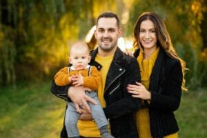 Szabadtéri családi fotózás Debrecenben. Egy boldog család őszi környezetben, napsütéses időben. Az apa tartja a kisfiát, aki a fényképezőgépre néz, miközben az anya mosolyogva áll mellette, kezét az apára helyezve. Mindhárom családtag sárga ruhákat visel, a háttérben zöld fák és napfény látszik.