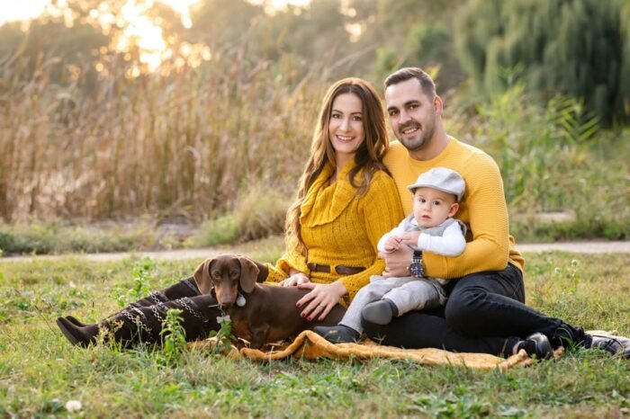 Egy boldog család ül a zöld fűben, mindannyian sárga pulóvereket viselnek. A kisfiú az anyukájával és apukájával van, egy kutyus pedig mellettük heverészve pihen. A háttérben a napsütéses táj és a fák láthatók, egy kellemes tavaszi nap hangulatát árasztva.