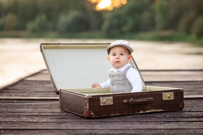 Szabatéri fotózás Debrecenben. Egy kisgyermek ült egy régi bőröndben, szép tavaszi környezetben, egy dombos tájon. Az apró fiú csinos kis öltönyben, sapkában és fehér pólóban pózol, miközben a háttérben a zöld fű és a napsütéses táj adja a varázslatos hangulatot a fotózásnak.