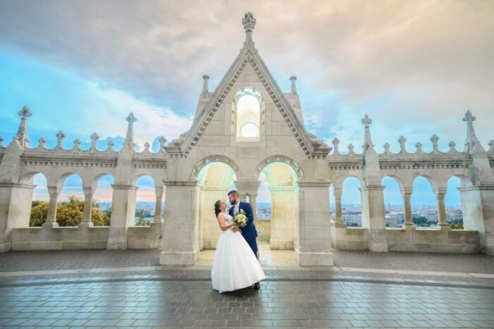 A menyasszony és a vőlegény romantikus pillanata a Halászbástyánál, Budapest híres épületénél. A kép hátterében lenyűgöző városi panoráma és a naplemente aranyfényei láthatóak, miközben a pár boldogan öleli egymást. A menyasszony fehér esküvői ruhája és virágcsokra ragyog a fényben, míg a vőlegény elegáns kék öltönyben mosolyog. A háttérben a Halászbástya jellegzetes tornyai és a város egyedülálló látképe adja a fotó különleges hangulatát.