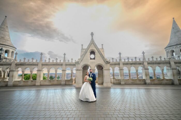 A menyasszony és a vőlegény romantikus pillanata a Halászbástyánál, Budapest szívében. A menyasszony fehér esküvői ruhájában, virágcsokorral a kezében, a vőlegény kék öltönyében, a háttérben Budapest lenyűgöző látképével és a Halászbástya mesés építészeti részleteivel. A kép különleges hangulatát a naplemente aranyfénye adja, miközben a pár egymásra figyel.