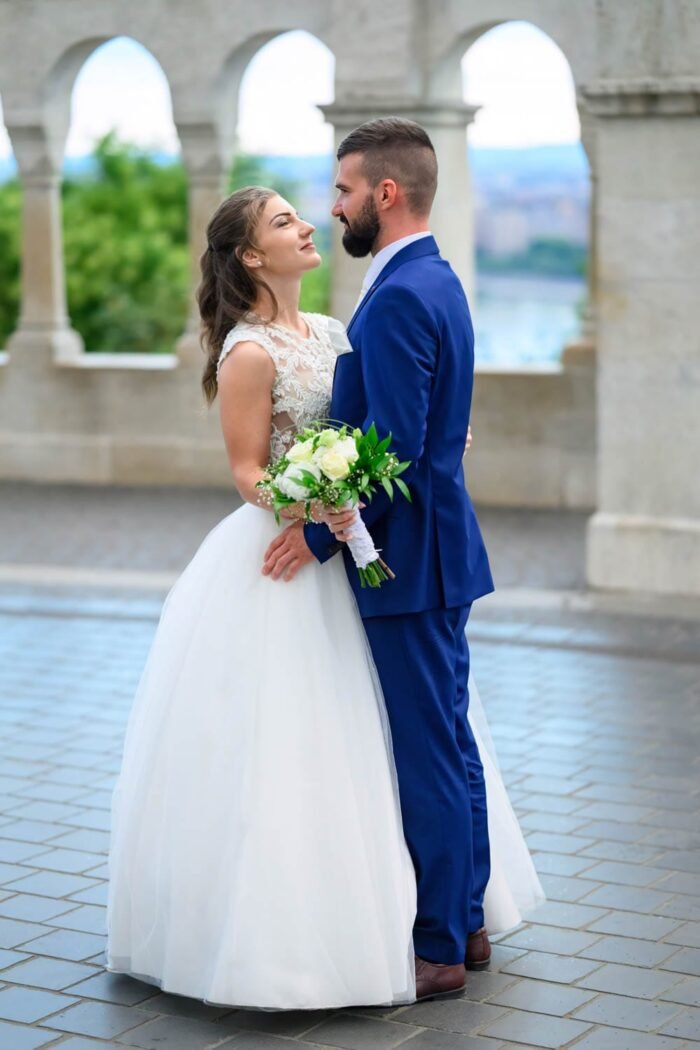 A menyasszony és a vőlegény romantikus pillanata a Halászbástya teraszán. A menyasszony gyönyörű fehér esküvői ruhájában, a vőlegény kék öltönyében állnak szemben egymással, mosolyogva és a menyasszony virágcsokorral a kezében. A háttérben Budapest lenyűgöző látképe, a Duna és a Parlament is látható, miközben az esküvői pár egymásra koncentrál.