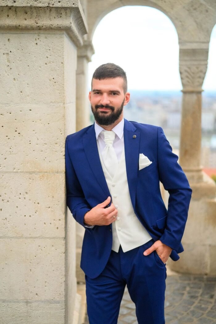 A vőlegény a Halászbástya egyik oszlopa mellett pózol, elegáns kék öltönyben és fehér nyakkendőben. A háttérben Budapest látképe és a város panorámája látható. A vőlegény mosolygós tekintettel áll, öltönyét finoman igazítja, miközben a történelmi épület környezete fokozza az esküvői pillanat különlegességét.