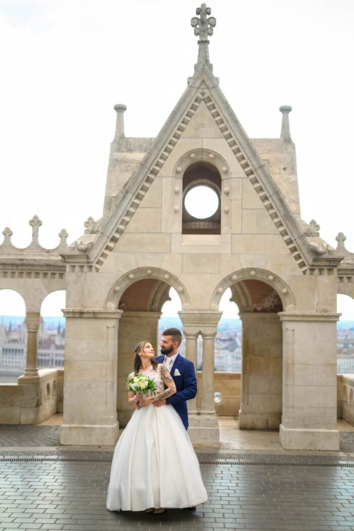 A friss házasok boldogan állnak egymás mellett a Halászbástyánál, miközben egymásra mosolyognak. A háttérben Budapest Parlamentje és a város panorámája látszik, a bástya építészeti elemei körülölelik a párt. A menyasszony gyönyörű esküvői ruhában, a vőlegény elegáns kék öltönyben, boldogan tartják egymást.
