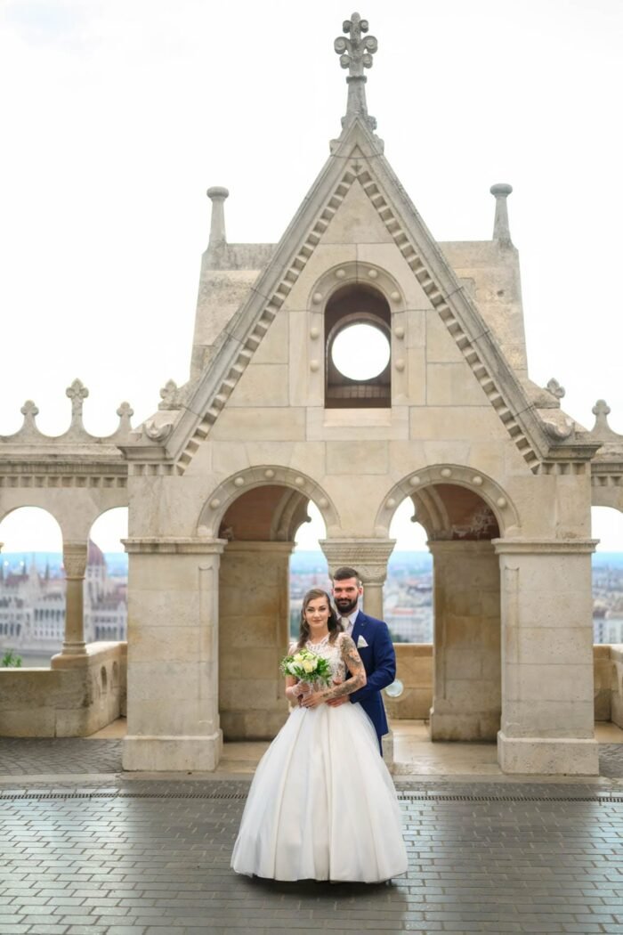 A menyasszony és vőlegény boldogan pózolnak a Halászbástyánál, háttérben Budapest Parlamentjével. A menyasszony gyönyörű esküvői ruhában és virágcsokorral, míg a vőlegény elegáns kék öltönyben áll a menyasszony mögött, keze a derekán. A történelmi bástyák és a város panorámája teszi még különlegesebbé a pillanatot.