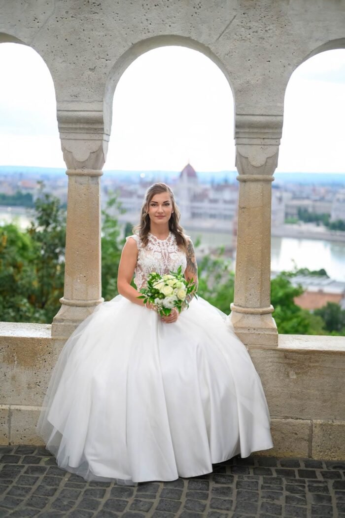 A menyasszony gyönyörű, hófehér esküvői ruhájában áll a Halászbástya teraszán, a Parlamenttel a háttérben. A menyasszony kezei között a virágcsokor, és a háttérben Budapest festői látképe látható.