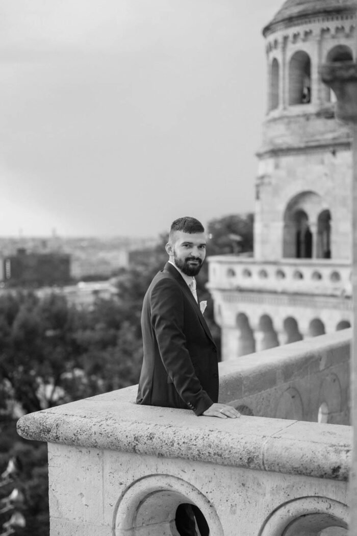 A vőlegény elegánsan pózol a Halászbástya teraszán, háttérben Budapest látképével és a monumentális építészeti részletekkel. A fekete-fehér fotó kiemeli a vőlegény karakterét és a város panorámáját.