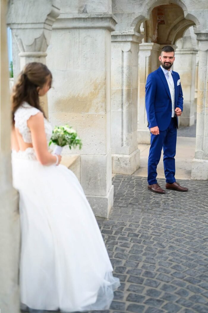 A menyasszony háttal állva, az esküvői csokorral a kezében, míg a vőlegény mosolyogva várja őt a Halászbástya egyik boltíve alatt. A háttérben a monumentális építészeti részletek és Budapest látképe.