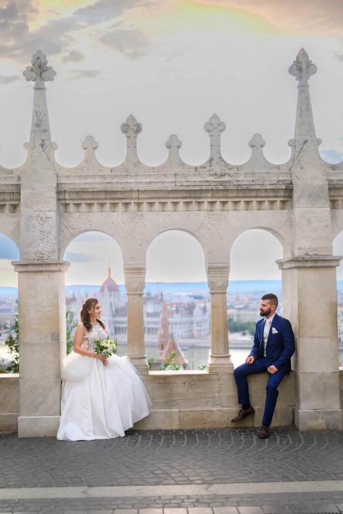 A menyasszony és a vőlegény romantikus pillanata a Halászbástya egyik boltíve alatt. A háttérben Budapest panorámája és a Parlament látható, miközben a menyasszony a férje mellett áll, ő pedig a bástya szélén pihen, mindketten boldogok és szeretetteljesek.