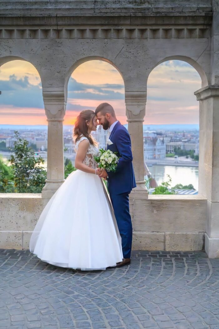 A házaspár romantikus pillanata a Halászbástya egyik boltíve alatt, a háttérben Budapest gyönyörű panorámájával és a naplementével. A menyasszony csodálatos ruhájában, a férj elegáns öltönyben, boldogan osztozva a szeretet pillanataiban.