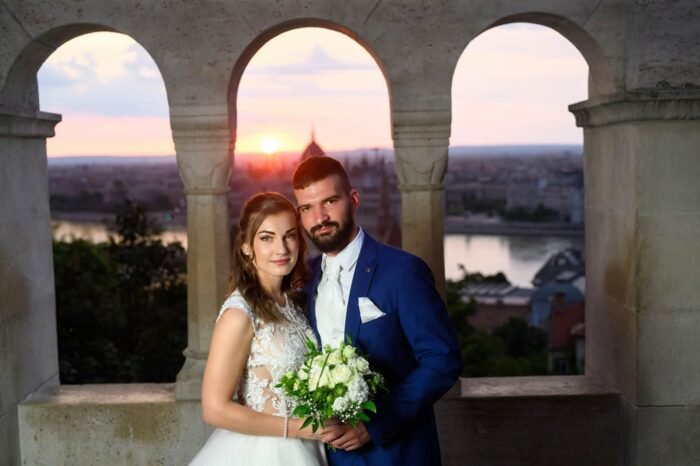 A házaspár a Halászbástya egyik ablakában, háttérben a napfelkeltével, Budapest gyönyörű panorámájával. A menyasszony csodálatos ruhájában, a férj elegáns öltönyben, csokorral a kezében, boldogan együtt.